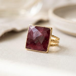 May include: A gold ring featuring a large, square-cut, faceted deep red gemstone set in a thin, textured gold frame. The band consists of three delicate, hammered gold wires that converge into a single shank at the base of the ring.