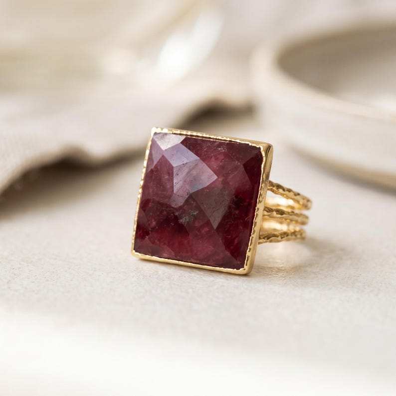 May include: A gold ring featuring a large, square-cut, faceted deep red gemstone set in a thin, textured gold frame. The band consists of three delicate, hammered gold wires that converge into a single shank at the base of the ring.
