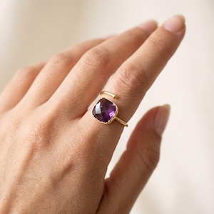 Könnte beinhalten: Ein goldfarbener offener Ring mit einem quadratisch geschliffenen, violetten Amethyst-Edelstein. Der Stein hat eine facettierte Oberfläche und das Band ist dünn mit einem kleinen abgerundeten Enddetail, was ein schlichtes Design ergibt.
