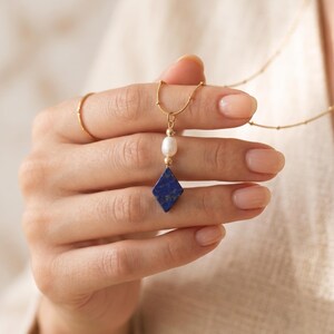 Könnte beinhalten: Eine goldene Halskette mit einem Anhänger aus einer kleinen weißen ovalen Perle und einem rautenförmigen blauen Lapislazuli-Stein. Die Kette besteht aus einer dünnen goldenen Kette mit kleinen goldenen Perlen und einem einfachen goldenen Ring.