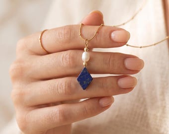 May include: A gold chain necklace featuring a pendant with a small white oval pearl and a diamond-shaped blue lapis lazuli stone. The necklace includes a thin gold chain with small gold beads spaced along the length and a simple gold ring.