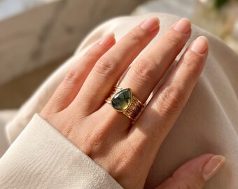 May include: A gold ring featuring a teardrop-shaped moss agate gemstone set in a wide, textured band. The metal has a polished finish, and the stone displays natural green inclusions within a translucent, pale grey and white crystalline structure.