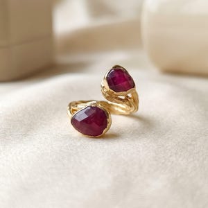 May include: A gold bypass ring featuring two faceted, pear-shaped deep red rubies. The band has a textured, organic metal finish that mimics the appearance of twisted branches or bark, connecting the two stones in an open design.
