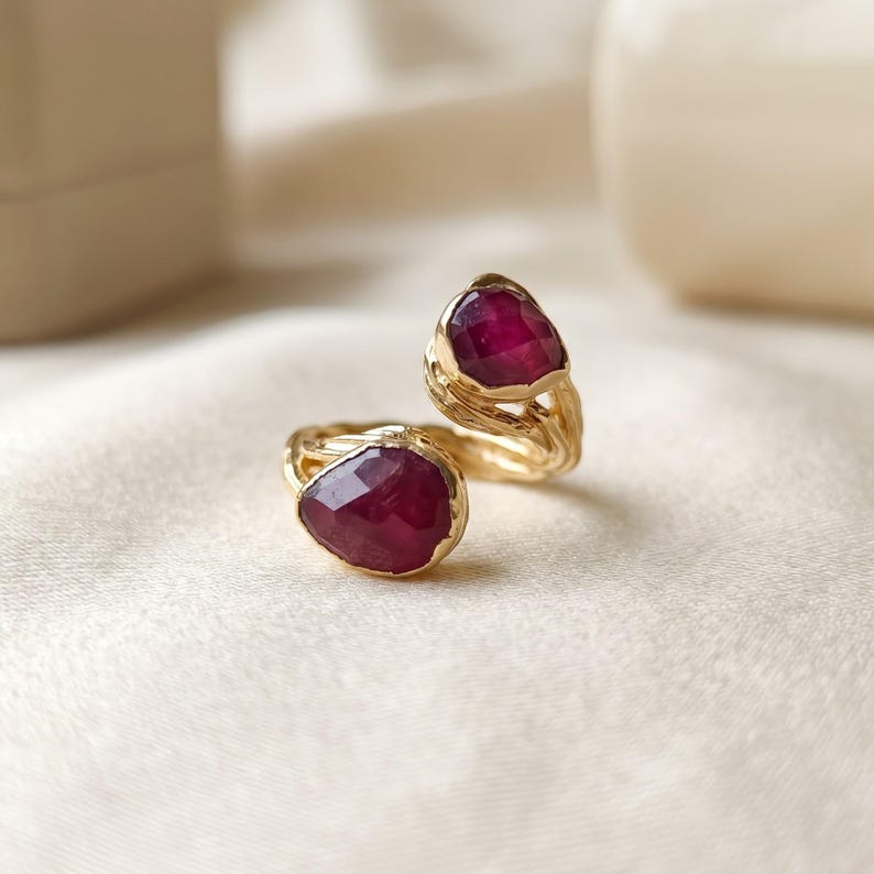 May include: A gold bypass ring featuring two faceted, pear-shaped deep red rubies. The band has a textured, organic metal finish that mimics the appearance of twisted branches or bark, connecting the two stones in an open design.