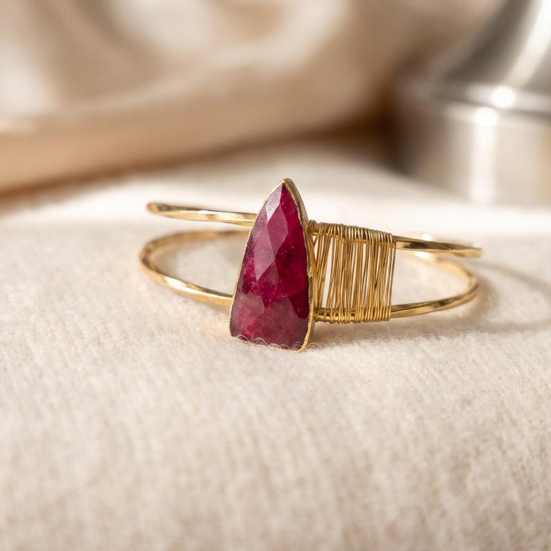 May include: A gold-toned cuff bracelet featuring a faceted, triangular-shaped deep red gemstone. The band is constructed from thin wire, with a section of the wire wrapped tightly around one side of the stone to secure it in place.