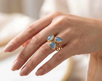 May include: A gold ring featuring three teardrop-shaped light blue stones arranged in a cluster. The band is thin and metallic, holding the three smooth, translucent cabochon-style stones securely in place on the finger with a polished finish.