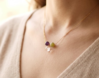 May include: A delicate gold-tone chain necklace featuring three small, round beads in purple, light green, and clear glass. The beads are suspended from a thin wire loop, creating a minimalist pendant design that rests against the collarbone.