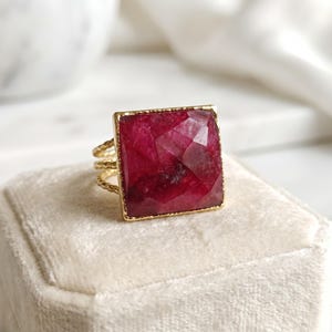 May include: A gold ring featuring a large, square, faceted deep red gemstone set in a thin, textured gold band. The stone has a visible geometric cut with dark and light crimson tones throughout its surface, held by a simple gold frame.
