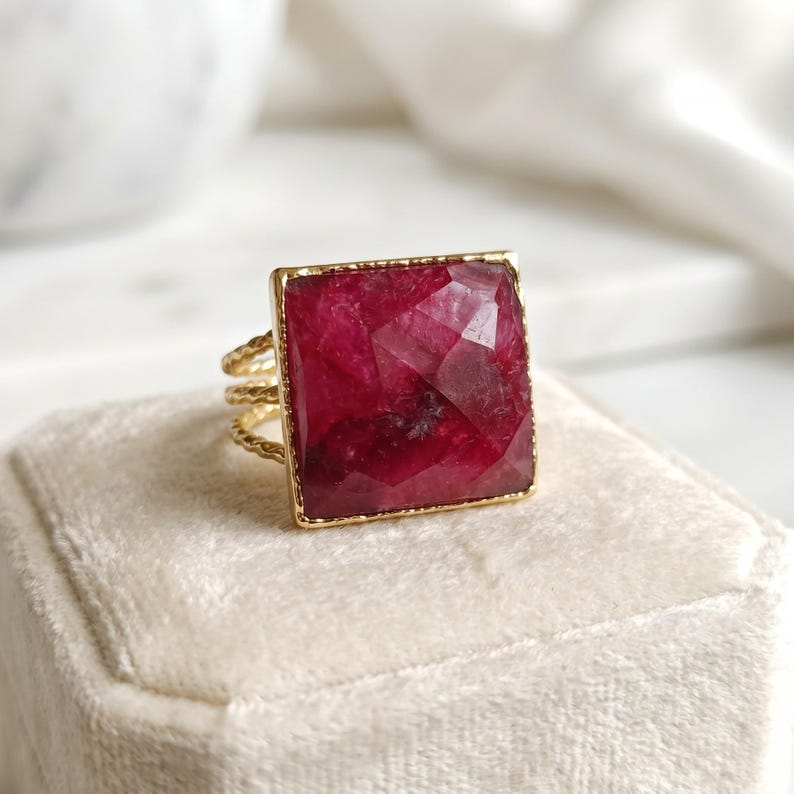 May include: A gold ring featuring a large, square, faceted deep red gemstone set in a thin, textured gold band. The stone has a visible geometric cut with dark and light crimson tones throughout its surface, held by a simple gold frame.