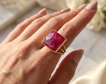 May include: A gold ring featuring a large, square-cut, faceted magenta gemstone. The band is constructed from three thin, twisted gold wires that converge to support the prominent, vibrant pink stone setting.