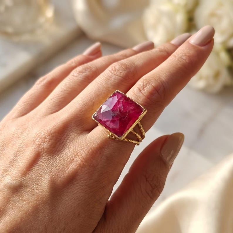 May include: A gold ring featuring a large, square-cut, faceted magenta gemstone. The band is constructed from three thin, twisted gold wires that converge to support the prominent, vibrant pink stone setting.