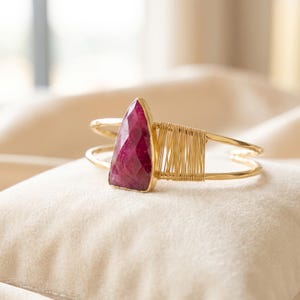 May include: A gold-toned cuff bracelet featuring a triangular, faceted deep pink gemstone set in a gold-coloured bezel. The bracelet band is constructed from two thin metal wires, with one side accented by a section of tightly coiled gold wire.