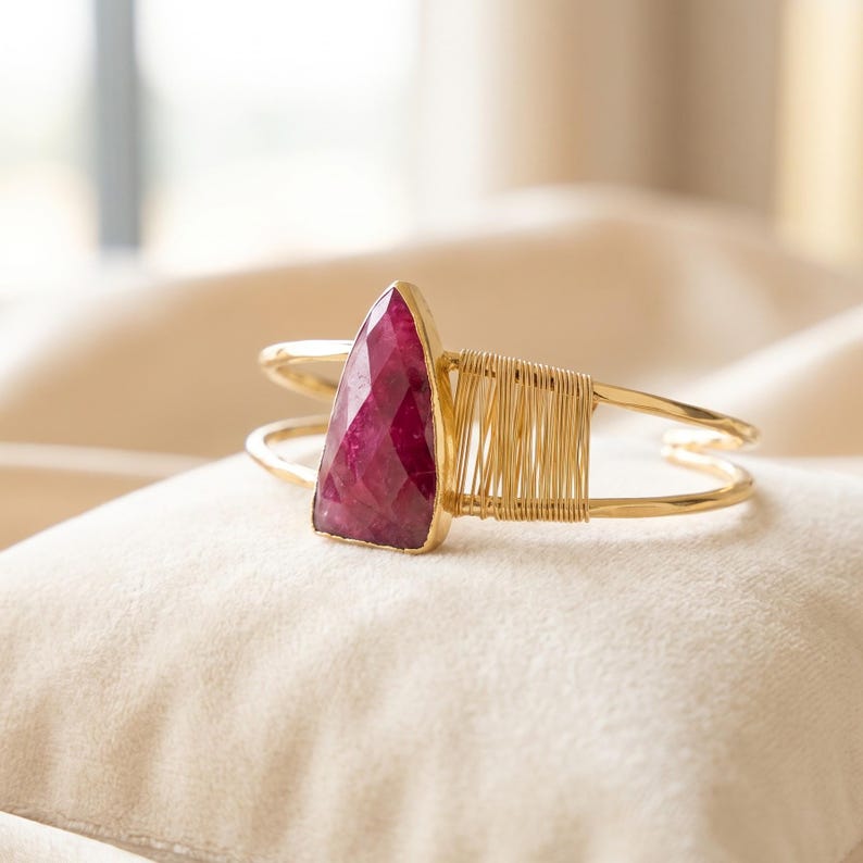 May include: A gold-toned cuff bracelet featuring a triangular, faceted deep pink gemstone set in a gold-coloured bezel. The bracelet band is constructed from two thin metal wires, with one side accented by a section of tightly coiled gold wire.