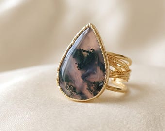 May include: A gold ring featuring a teardrop-shaped moss agate gemstone with dark green dendritic inclusions. The stone is set in a smooth gold bezel, and the band is constructed from multiple thin, layered gold wires that wrap around the finger.
