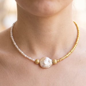 May include: A necklace featuring a central large, irregular white pearl flanked by two textured gold-toned spherical beads. One side consists of small white seed pearls, while the other side features a strand of small, flat gold-toned disc beads.