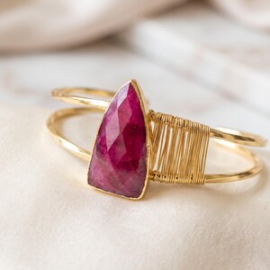 May include: A gold-toned cuff bracelet featuring a faceted, triangular-shaped deep pink gemstone. The band is constructed from two parallel wires, with one side accented by a decorative section of tightly coiled gold-toned wire wrapping.