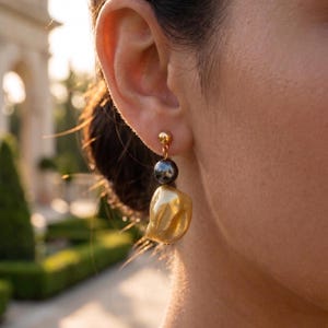 May include: A gold stud earring featuring a small gold sphere at the top, a dark grey spherical bead in the centre, and a larger, irregular, textured gold drop pendant at the bottom, all suspended from a gold post and backing mechanism.
