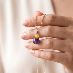 May include: A gold chain necklace featuring a pendant with three stacked beads: a small dark purple bead, a medium yellow bead, and a large faceted dark purple bead at the bottom. The chain has small gold accents spaced along its length.