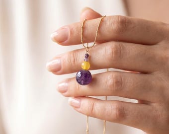 Puede incluir: Un collar de cadena dorada con un colgante de tres cuentas apiladas: una cuenta pequeña de color morado oscuro, una cuenta mediana amarilla y una cuenta grande facetada de color morado oscuro en la parte inferior.