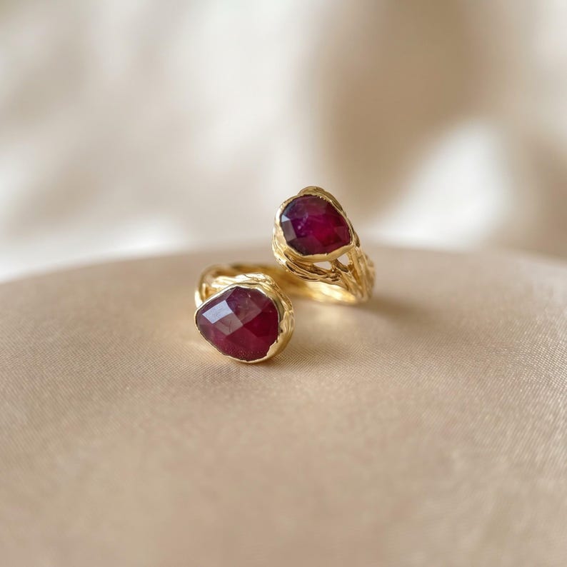 May include: A gold open ring featuring two faceted, deep red, pear-shaped gemstones. The band has a textured, organic, branch-like appearance, with the two stones positioned at the open ends of the ring, facing each other.