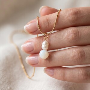 May include: A gold-toned chain necklace featuring a delicate pendant composed of one small, oval-shaped white pearl stacked above a single, larger, smooth, round white opal-like bead, suspended from a thin, textured gold-coloured metal chain.