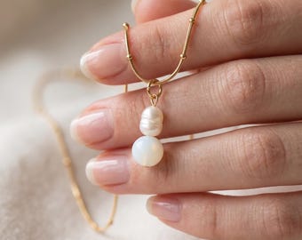 May include: A gold-toned chain necklace featuring a delicate pendant composed of one small, oval-shaped white pearl stacked above a single, larger, smooth, round white opal-like bead, suspended from a thin, textured gold-colored metal chain.