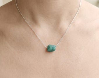 May include: A delicate silver chain necklace featuring a single, raw, uncut emerald-green gemstone pendant. The stone has a rough, natural texture and is suspended centrally on the thin, polished metallic link chain.