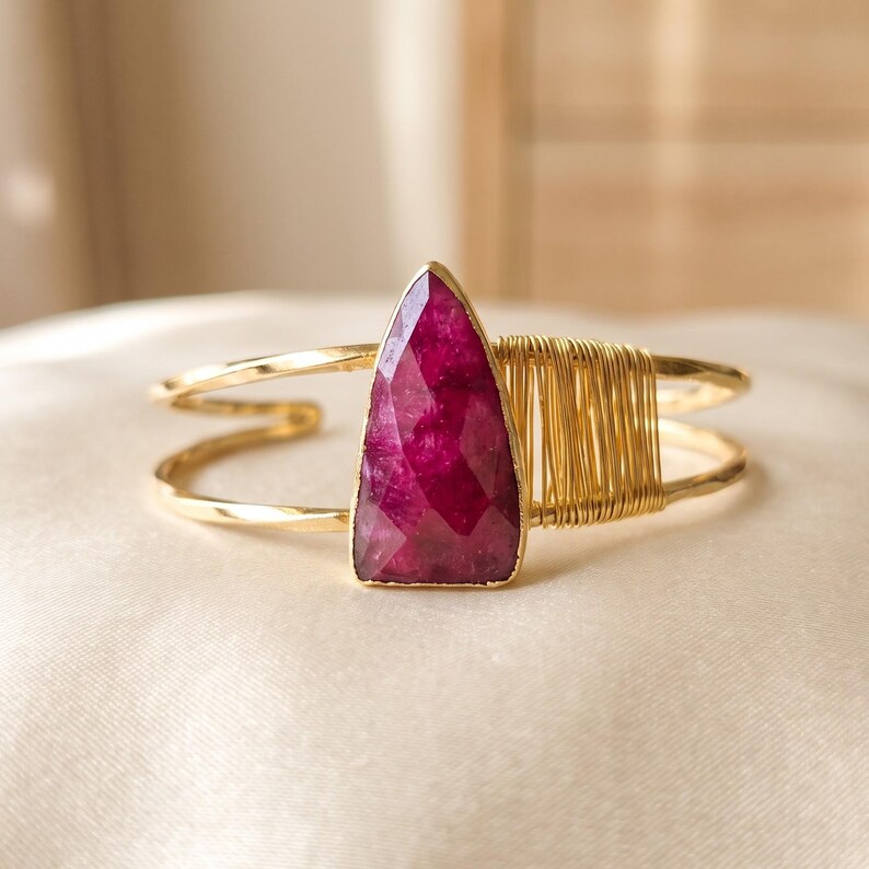 May include: A gold-toned cuff bracelet featuring a central, triangular, faceted deep pink gemstone. The band is constructed from two parallel wires, with one side accented by a decorative wrapping of thin gold-coloured wire.
