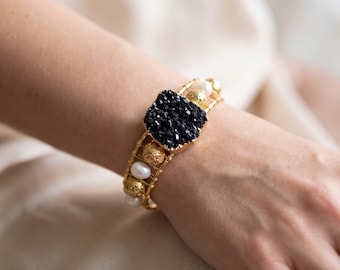 May include: A gold wire cuff bracelet featuring a central square black druzy stone, accented with alternating white pearls and textured gold-tone spherical beads. The bracelet is constructed with a multi-strand wire design that wraps around the wrist.