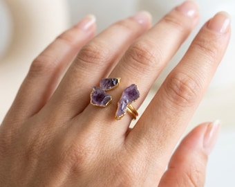 May include: A gold-toned open ring featuring three raw, purple amethyst stones. The ring has a split design with two smaller amethyst pieces on one side and a single, elongated amethyst piece on the other, all set in gold-colored metal.