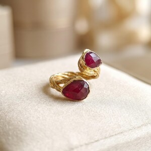 May include: A gold open-style ring featuring two faceted, pear-shaped deep red rubies set at each end. The band has a textured, twisted metal finish that resembles organic vine-like strands, connecting the two gemstone settings.