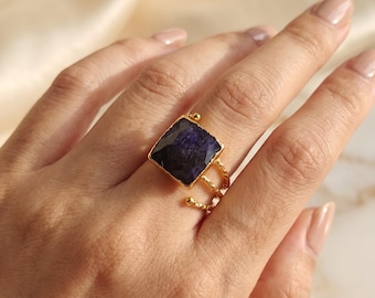 May include: A gold ring featuring a large, square-cut, deep purple gemstone set in a textured metal frame. The band consists of three thin, twisted gold wires that converge into a single shank, providing a layered appearance on the finger.