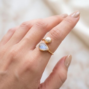 Könnte beinhalten: Ein goldfarbener offener Ring mit einer runden weißen Perle und einem birnenförmigen, irisierenden Mondstein. Das Band ist dünn und metallisch, so gestaltet, dass es sich teilweise um den Finger legt und zwei Steine im Bypass-Stil zeigt.