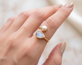 May include: A gold-toned open ring featuring a round white pearl and a pear-shaped iridescent moonstone. The band is thin and metallic, designed to wrap partially around the finger, showcasing two distinct gemstone settings in a bypass style.