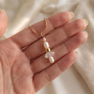 May include: A gold-toned chain necklace featuring a pendant composed of two small white freshwater pearls, a central smooth gold-toned cylindrical bead, and a clear, irregularly shaped raw crystal fragment positioned between the pearls.