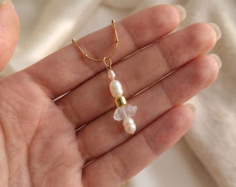 May include: A gold-toned chain necklace featuring a pendant composed of two small white freshwater pearls, a central smooth gold-toned cylindrical bead, and a clear, irregularly shaped raw crystal fragment positioned between the pearls.