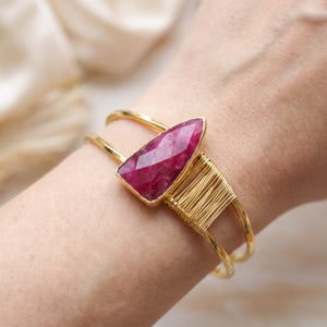 May include: A gold-toned cuff bracelet featuring a faceted, triangular-shaped deep pink gemstone. The band is constructed from two thin metal wires, with one side accented by a decorative section of tightly coiled gold-coloured wire.