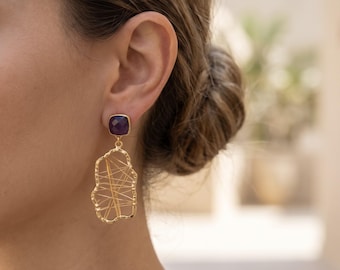 May include: A gold-tone earring featuring a square-cut purple amethyst stud at the top, connected to a large, irregular, open-frame gold-tone pendant with a delicate web of thin gold-tone wire crisscrossing the center of the shape.