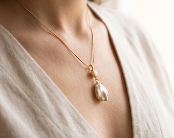 May include: A gold-toned chain necklace featuring a pendant with a small round white pearl positioned above a larger, irregular-shaped baroque pearl. Both pearls are set in gold-toned metal, creating a layered drop design on a thin metallic chain.