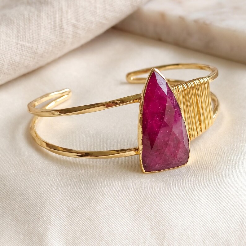 May include: A gold-toned cuff bracelet featuring a large, faceted, teardrop-shaped magenta gemstone. The band is constructed from two parallel wires, with one side accented by a series of tightly wrapped gold-toned wire coils.