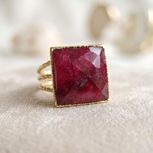 May include: A gold ring featuring a large, square-cut, faceted deep red gemstone set in a thin gold frame. The band of the ring is composed of three delicate, textured, wavy gold wires that connect to the base of the square setting.