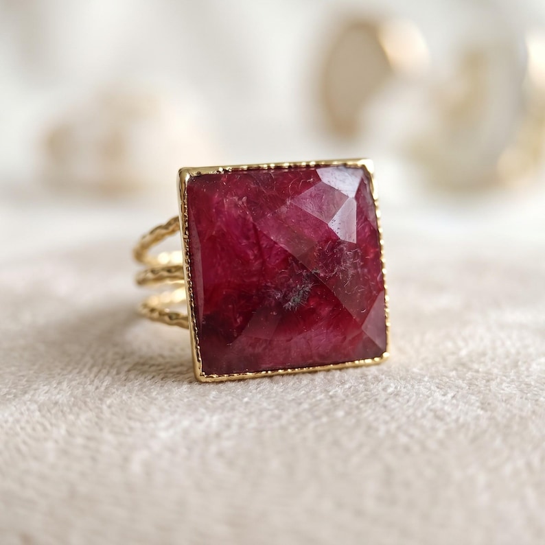 May include: A gold ring featuring a large, square-cut, faceted deep red gemstone set in a thin gold frame. The band of the ring is composed of three delicate, textured, wavy gold wires that connect to the base of the square setting.
