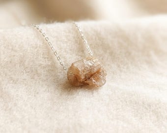 May include: A delicate silver chain necklace featuring a raw, light brown, translucent crystal pendant. The stone has a rough, multifaceted texture and is suspended from a thin, polished metallic link chain that catches the light.