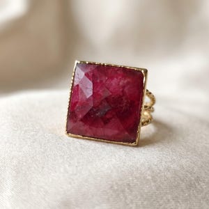 May include: A gold ring featuring a large, square-cut, faceted deep red gemstone set in a thin, textured gold frame. The band of the ring is composed of multiple thin, twisted gold wires that connect to the base of the square setting.