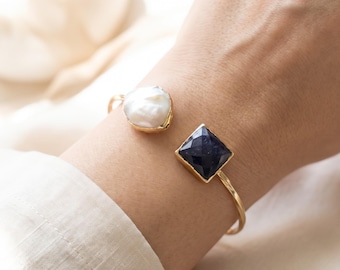 May include: A gold-toned open cuff bracelet featuring a white irregular baroque pearl on one end and a square, faceted dark blue gemstone on the other. The metal band is thin and polished, creating a contrast between the organic pearl and the geometric stone.