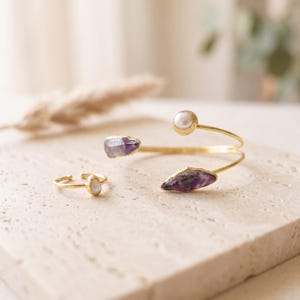 Può includere: Un bracciale aperto color oro e un anello aperto abbinato. Il bracciale presenta una perla bianca rotonda a un'estremità e un cristallo di ametista viola grezzo all'altra. L'anello è decorato con una piccola perla bianca rotonda.