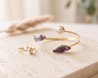 May include: A gold-toned open cuff bracelet and a matching open ring. The bracelet features a round white pearl at one end and a raw, purple amethyst crystal at the other. The ring is adorned with a single, small, round white pearl.