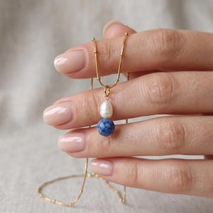 May include: A gold chain necklace featuring a delicate beaded texture along the links. The pendant consists of a single white oval freshwater pearl positioned directly above a round, mottled blue sodalite gemstone bead on a gold jump ring.