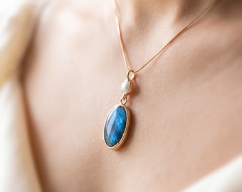 May include: A gold chain necklace featuring a pendant with an oval-shaped blue labradorite gemstone and a small, round white pearl accent. The pendant is suspended from a delicate gold-toned metal chain that rests against the collarbone area.