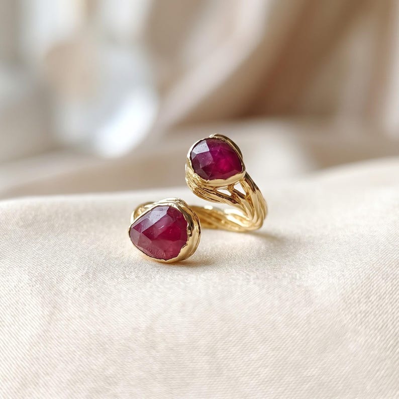 May include: A gold open-style ring featuring two faceted, deep red ruby-colored stones. The band has a textured, organic, branch-like appearance that connects the two stones at opposite ends of the open design, creating a bypass ring silhouette.